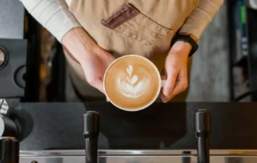 Imagem de um barista vestindo um avental marrom segura uma xícara de café com leite com arte em forma de coração, visto de cima do balcão de uma cafeteria, para ilustrar matéria sobre a Confeitaria Colombo (RJ).