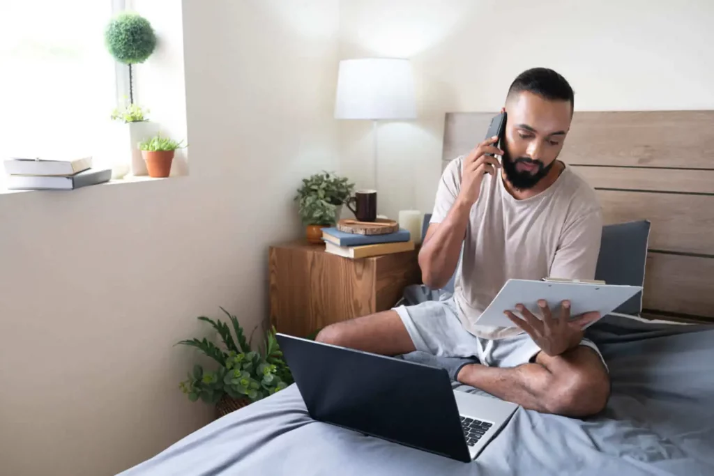 Imagem de um homem sentado de pernas cruzadas em uma cama com um notebook à sua frente, falando ao telefone e lendo uma prancheta. Ele está em um quarto bem iluminado com plantas, livros e uma lâmpada em uma mesa de cabeceira de madeira.