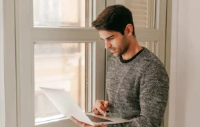 Imagem de um homem de cabelos escuros, vestindo um suéter cinza, está ao lado de uma janela, segurando e usando um notebook, parecendo concentrado na tela em um ambiente interno claro.