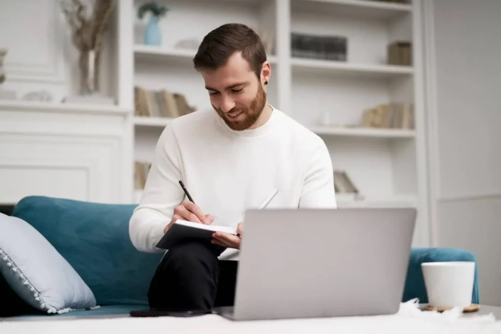 Imagem de um homem com barba sentado em um sofá, sorrindo enquanto escreve em um caderno, para ilustrar matéria que responde a pergunta: posso desistir do consórcio? Ele tem um notebook à sua frente e uma caneca branca por perto, em uma sala de estar aconchegante com prateleiras de livros ao fundo.