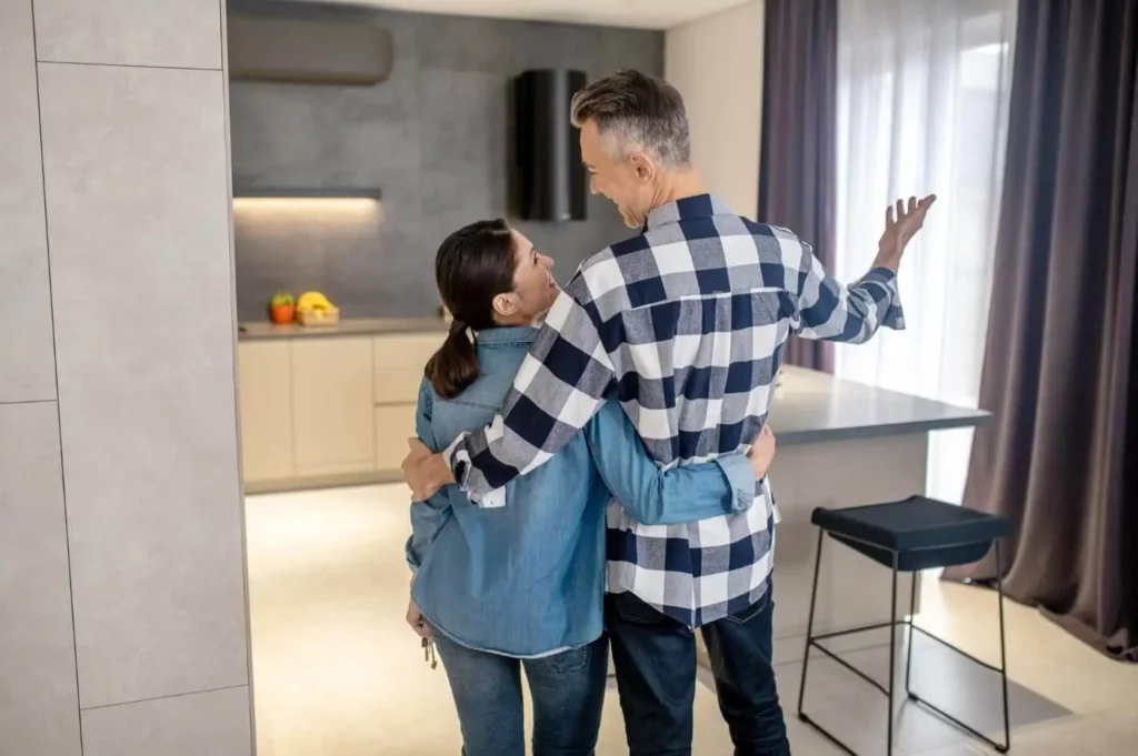 Imagem de um casal de braços dados em uma cozinha moderna, sorrindo um para o outro. O homem faz um gesto em direção ao cômodo, enquanto a mulher segura as chaves e olha para ele.