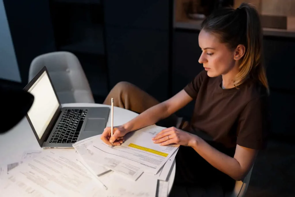 Imagem de uma jovem mulher sentada em uma mesa com um notebook e papéis espalhados, preenchendo formulários com uma caneta. Ela parece concentrada e está trabalhando em uma sala pouco iluminada.