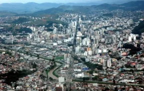 Vista aérea da cidade de Juiz de Fora, com inúmeros prédios altos, um rio que a atravessa pelo meio, ruas e casas, tendo como pano de fundo colinas e montanhas verdes.