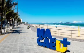 Um calçadão ensolarado à beira-mar, com palmeiras, uma calçada larga e um grande letreiro azul e amarelo com a palavra “Macaé” em primeiro plano. A praia arenosa e o mar se estendem até o horizonte sob um céu azul límpido.