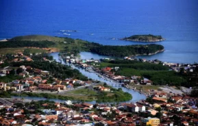 Vista aérea de uma parte da orla de Rio das Ostras, com prédios de telhados vermelhos, um rio serpenteando pelo centro, áreas verdes exuberantes, pontes e pequenas ilhas cercadas pelas águas azuis do oceano ao fundo.