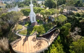 Vista aérea de um monumento branco com uma estátua, cercado por árvores, caminhos e um mirante de madeira em forma de curva, em um parque verdejante, com a cidade visível ao fundo.