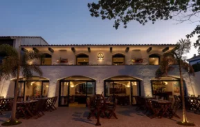 Um restaurante de dois andares com iluminação aconchegante, paredes de estuque branco, portas em arco, mesas e cadeiras de madeira ao ar livre e palmeiras emoldurando a entrada ao anoitecer.