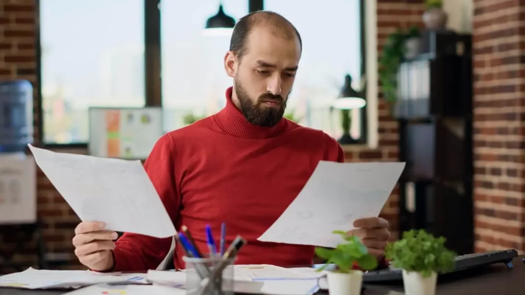 Imagem de um homem vestindo uma blusa de gola alta vermelha está sentado à mesa, segurando duas folhas de papel, com ar de concentração. A mesa está cheia de papéis, canetas e pequenos vasos de plantas. Ao fundo, vêem-se prateleiras e grandes janelas em um escritório moderno.