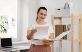 Imagem de uma mulher em um escritório, sorrindo enquanto folheia documentos em uma pasta. Ela está vestindo uma camisa branca e calças marrom-claro, com prateleiras e um laptop visíveis ao fundo.