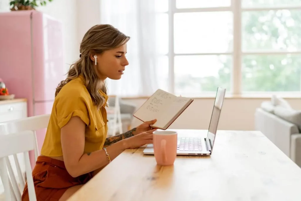 Imagem de uma mulher usando fones de ouvido, sentada à mesa com um laptop, segurando um caderno aberto e uma caneta. Há uma caneca rosa à sua frente, e ela parece estar trabalhando ou estudando em uma sala bem iluminada.