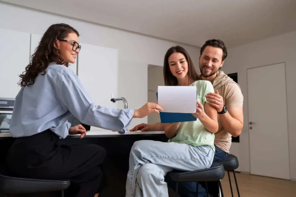 Imagem de um mulher de óculos mostrando alguns documentos a um casal sorridente que está à frente. O casal parece feliz e envolvido, examinando os papéis juntos em uma cozinha moderna e bem iluminada.