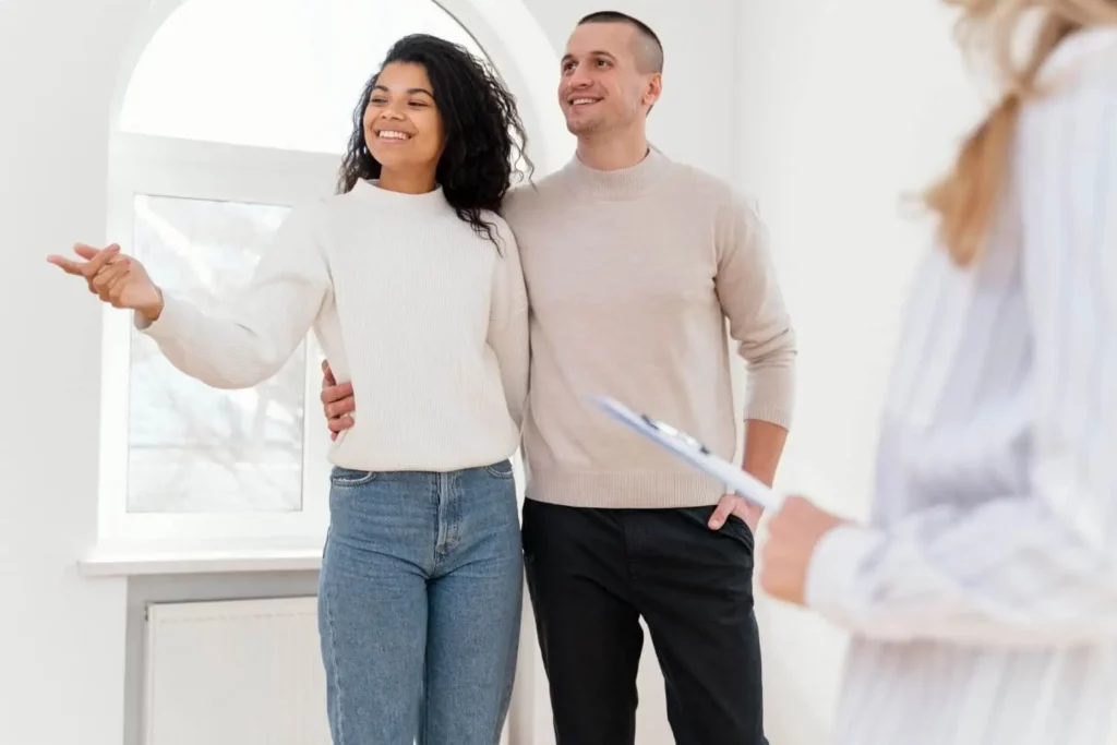 Imagem de um casal sorridente está junto em uma sala bem iluminada, com uma das pessoas apontando para a frente. Outra pessoa, segurando uma prancheta, aparece parcialmente em primeiro plano, possivelmente mostrando o local ao casal.