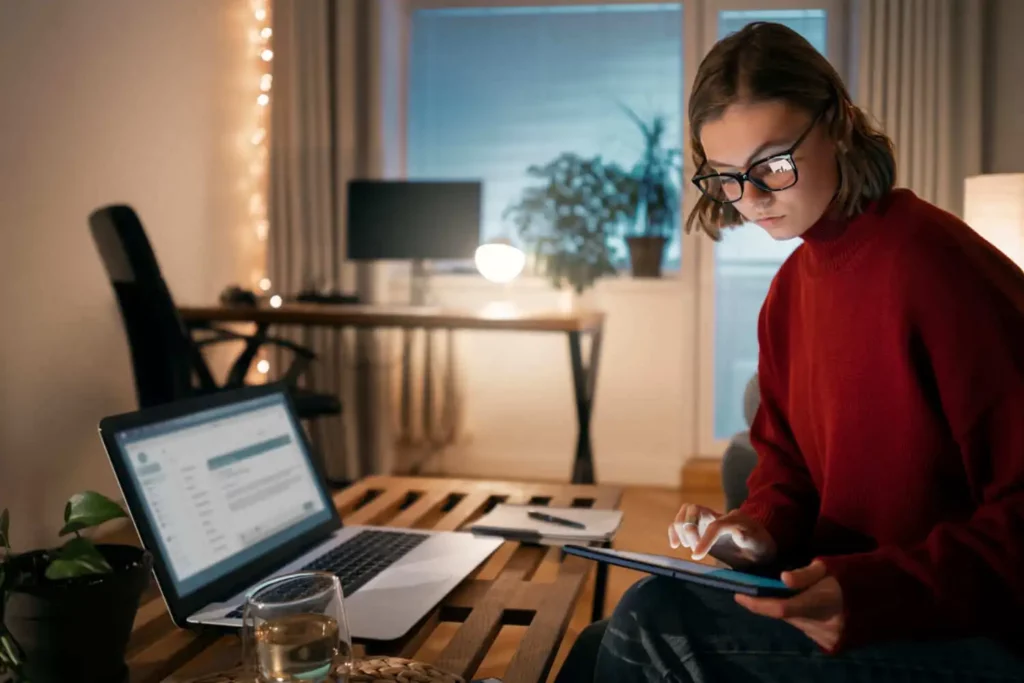 Imagem de uma mulher de óculos e suéter vermelho trabalha em uma mesa de madeira com um laptop e um tablet em uma sala aconchegante, com iluminação suave, plantas e luzes decorativas ao fundo.