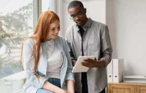 Imagem de uma mulher de cabelos ruivos longos está sentada em uma mesa, sorrindo, enquanto um homem de óculos está ao lado dela, segurando e mostrando algo a ela em um tablet. Eles parecem estar tendo uma conversa amigável em um ambiente de escritório.