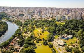 Vista aérea de uma cidade com muitos prédios altos, um rio sinuoso e um grande parque verde repleto de árvores, espaços abertos e um lago.