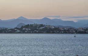 Um mar calmo com um veleiro solitário, uma cidade litorânea situada em uma pequena colina e camadas de montanhas azuis ao fundo, sob um céu em tons pastéis ao entardecer.
