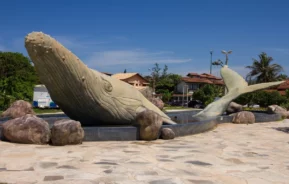 Grande escultura de uma baleia parcialmente submersa na fonte de uma praça, cercada por rochas. No fundo, avistam-se casas, árvores e um céu azul límpido.