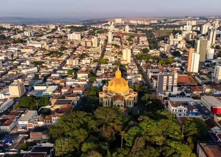 Vista aérea de uma cidade com um grande edifício de cúpula amarela cercado por árvores em primeiro plano e muitos prédios residenciais ao fundo, sob um céu limpo.
