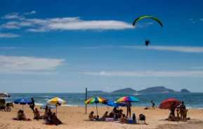 As pessoas relaxam sob guarda-sóis coloridos em uma praia de areia à beira-mar, enquanto, no céu azul acima, alguém pratica parapente. Ilhas distantes são visíveis no horizonte, sob nuvens esparsas.