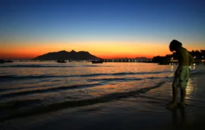 Uma pessoa caminha pela orla ao pôr do sol, com ondas suaves e montanhas distantes recortadas contra um céu alaranjado e azul.