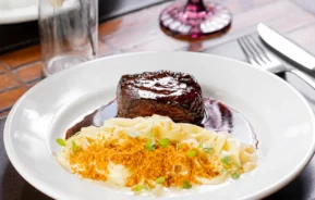 Um prato branco com um bife glaceado, fettuccine cremoso coberto com migalhas crocantes e ervas, servido com um molho escuro, colocado sobre a mesa de um restaurante com um garfo, uma faca e um copo roxo ao fundo. O prato é do restaurante Assunta, em Juiz de Fora