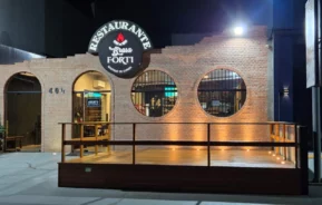 Um restaurante de alvenaria à noite, com janelas circulares, um pátio de madeira e uma placa iluminada com os dizeres “Restaurante Brasa Forti”, um dos principais Restaurantes em Limeira. A porta de entrada está aberta, e as luzes iluminam a fachada.