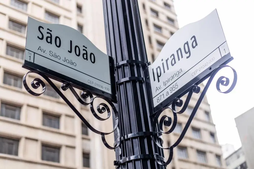 Imagem de placas de sinalização no cruzamento da Avenida São João com a Avenida Ipiranga, fixadas em um poste preto, com prédios altos ao fundo.