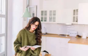 Imagem de uma mulher de cabelos castanhos e óculos lendo um livro em uma cozinha bem iluminada e moderna para ilustrar matéria sobre como funciona a contemplação no consórcio. Ela veste um suéter verde largo e está sentada perto de uma bancada de madeira, com uma caneca e fones de ouvido.
