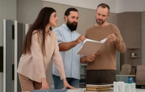 Imagem de três pessoas em volta de uma mesa em um escritório, examinando documentos juntas. Um homem segura e aponta para os papéis, enquanto o outro homem e uma mulher observam atentamente. Há livros e materiais de escritório sobre a mesa.