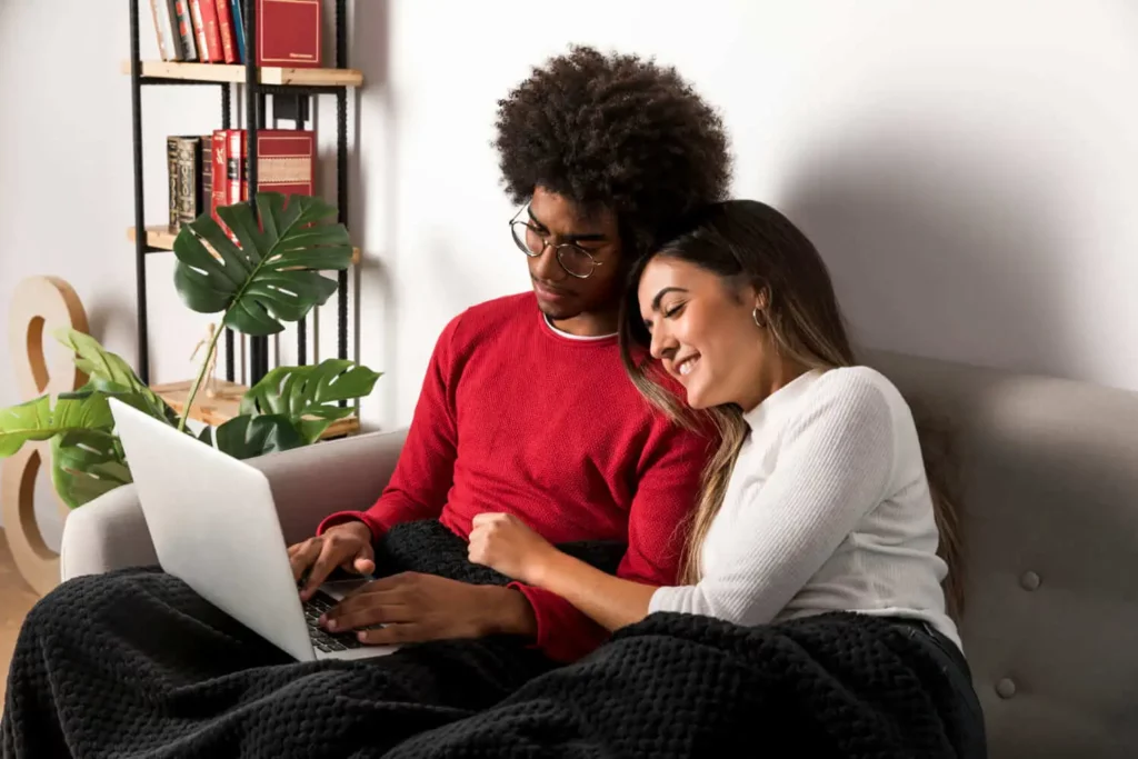 Imagem de um casal sentado em um sofá, coberto por um cobertor preto, olhando juntos para um notebook e sorrindo, para ilustrar matéria sobre o que é a taxa de administração no consórcio. Há livros e uma planta em uma estante ao fundo.