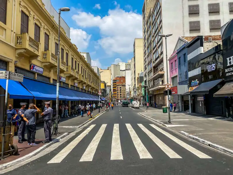 Imagem da Rua 25 de Março, em São Paulo, mostra uma faixa de pedestres, ladeada por prédios amarelos e azuis. As pessoas estão nas calçadas, sob toldos azuis. O céu está parcialmente nublado.