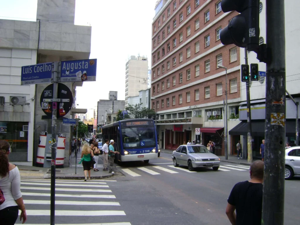 Imagem da Rua Augusta, em São Paulo, mostra pessoas atravessando a rua, um ônibus e um carro em uma faixa de pedestres. As placas de rua indicam “Luis Coelho” e “Augusta”. Edifícios altos, fachadas de lojas e semáforos são visíveis.(Foto: Wikimedia Commons)