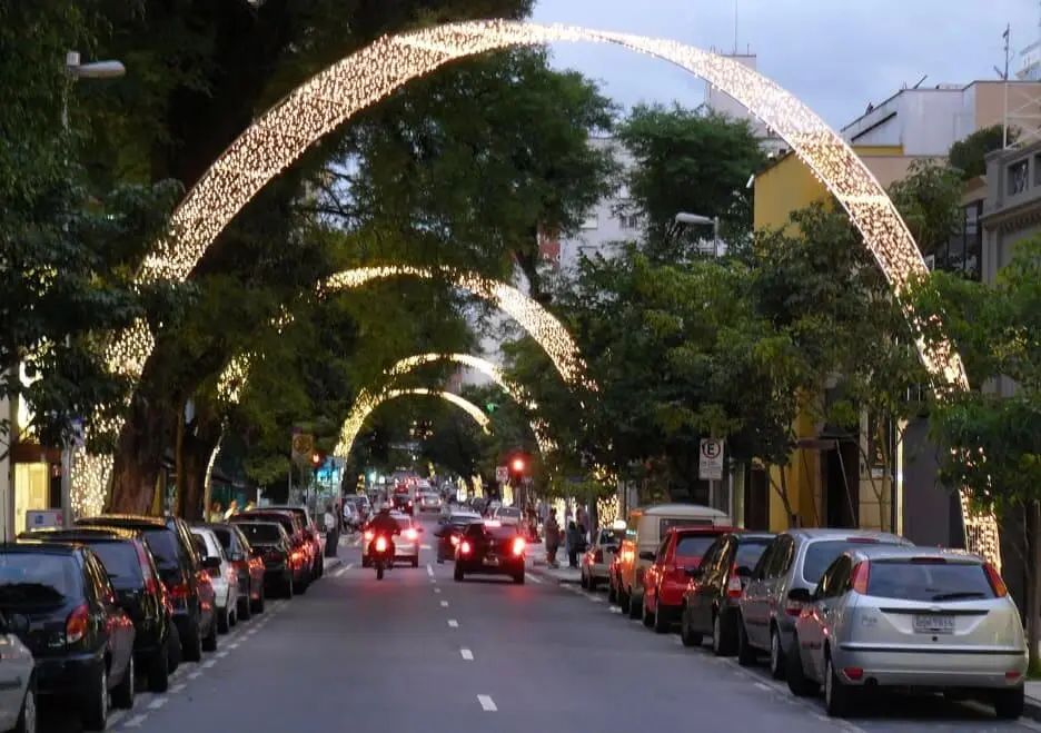 Imagem da Rua Oscar Freire, em São Paulo, mostra carros estacionados e árvores, decorada com grandes arcos iluminados de luzes que se estendem sobre a via. Pessoas e motocicletas circulam pela rua sob os arcos brilhantes ao anoitecer. (Foto: Wikimedia Commons)