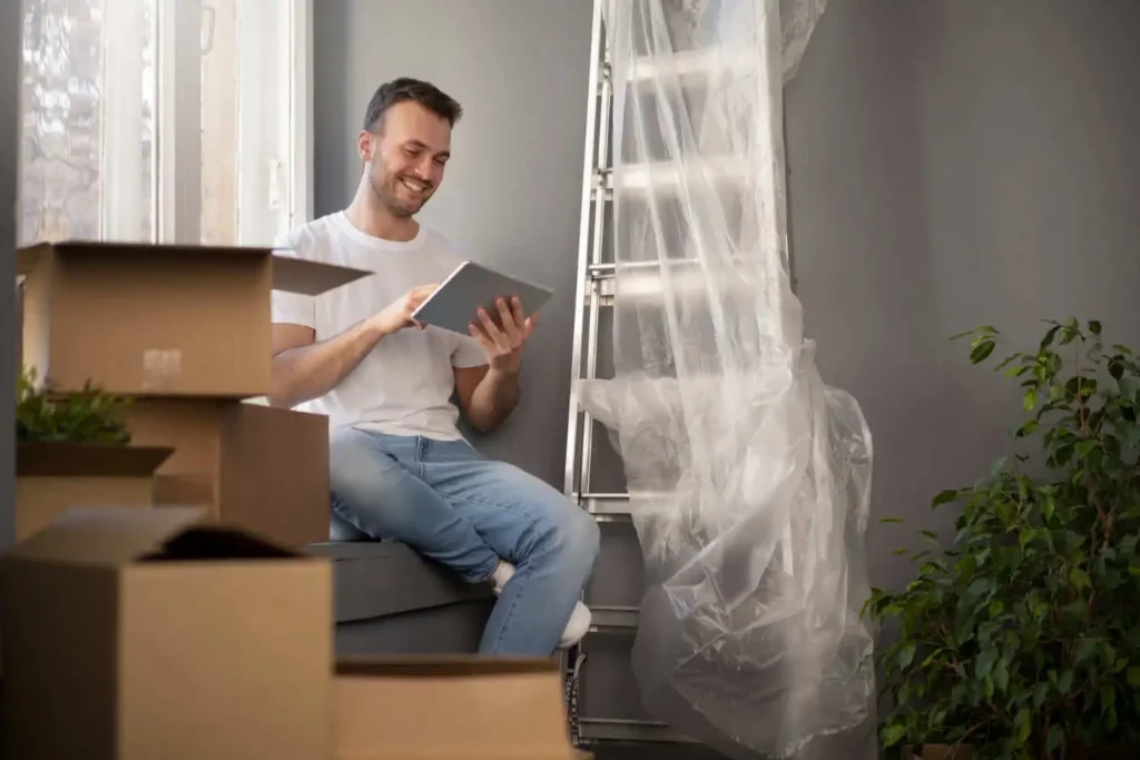 Imagem de um homem vestindo camiseta branca e jeans está sentado perto de caixas de mudança, sorrindo enquanto usa um tablet, para ilustrar matéria sobre a taxa do consórcio. Uma escada coberta com plástico e um vaso de planta estão perto.