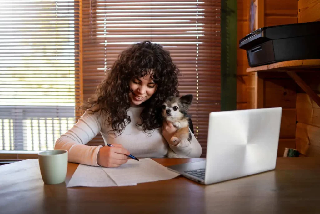 Imagem de uma mulher de cabelos cacheados escrevendo em um papel, sentada em uma mesa de madeira onde há um notebook, uma xícara e uma impressora, para ilustrar matéria sobre como calcular a taxa de administração do consórcio. Ela sorri enquanto segura um cachorrinho. A luz do sol entra pelas persianas atrás dela.