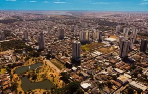 Vista aérea de uma cidade com vários prédios altos, um parque com lagos em primeiro plano e áreas urbanas que se estendem até o horizonte sob um céu azul.