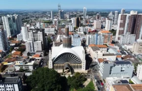 Vista aérea de uma grande catedral moderna com um teto abobadado único, cercada por prédios altos e ruas da cidade em um dia de céu limpo.