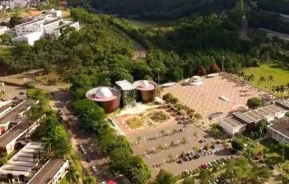 Vista aérea de um complexo de edifícios modernos com telhados arredondados, um estacionamento e uma grande praça aberta, cercada por árvores e vegetação, com colinas ao fundo.
