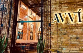 A entrada de um restaurante com paredes de tijolos e iluminação aconchegante, com o nome “AVVÍ” em letras iluminadas, plantas altas ao lado da porta e uma vista das mesas e cadeiras de madeira no interior.