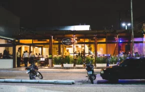 Vista da rua de um restaurante bem iluminado à noite, com pessoas jantando no interior, visíveis através das grandes janelas. Três motocicletas e parte de um carro estão estacionados em frente à entrada.