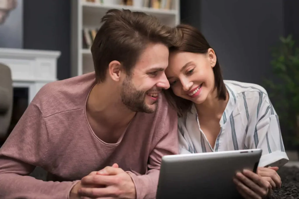 Imagem de um casal sorridente deitado bem próximo um do outro no chão, olhando para um tablet. Ao fundo, podem-se ver estantes de livros e uma lareira branca.