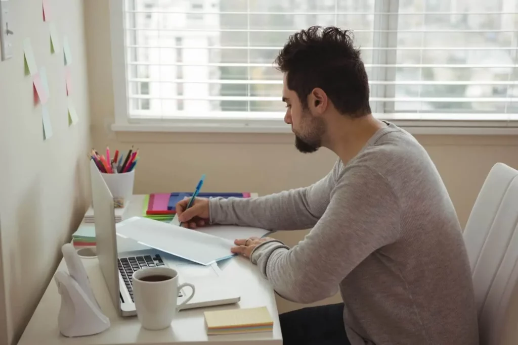 Imagem de um homem sentado à mesa, usando um notebook e escrevendo em um papel. Ao lado, há uma caneca de café, blocos de notas e canetas coloridas. Há post-its na parede e, ao fundo, uma janela com persianas.
