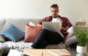 Imagem de um homem sentado em um sofá, trabalhando em um notebook, para ilustrar matéria que explica como funciona o consórcio com a Selic alta. Há almofadas coloridas, uma caixa de lenços, um vaso de planta e cadernos sobre a mesa de centro de vidro à sua frente.