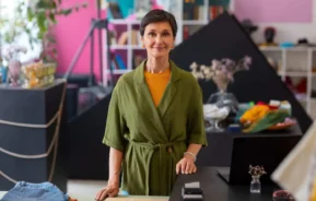 Imagem de uma mulher de cabelo curto e escuro, vestindo um vestido verde tipo envoltório e uma blusa amarela, de pé, sorrindo, atrás de uma mesa em um espaço de trabalho colorido e moderno, com plantas e objetos ao seu redor.