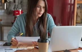 Imagem de uma mulher sentada à mesa, com alguns papéis e uma caneta na mão, olhando para a tela de um laptop. Perto dela há uma xícara de café, óculos e um celular. Ela parece concentrada e está em um ambiente aconchegante em casa.