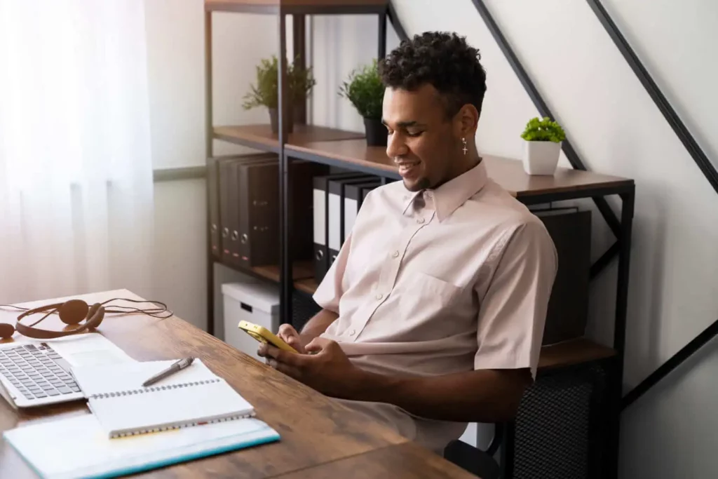 Um homem sentado em frente a uma mesa, com um notebook, um caderno e fones de ouvido, sorrindo enquanto olha para o celular. Ele está em um escritório bem iluminado e moderno, com estantes e plantas ao fundo.