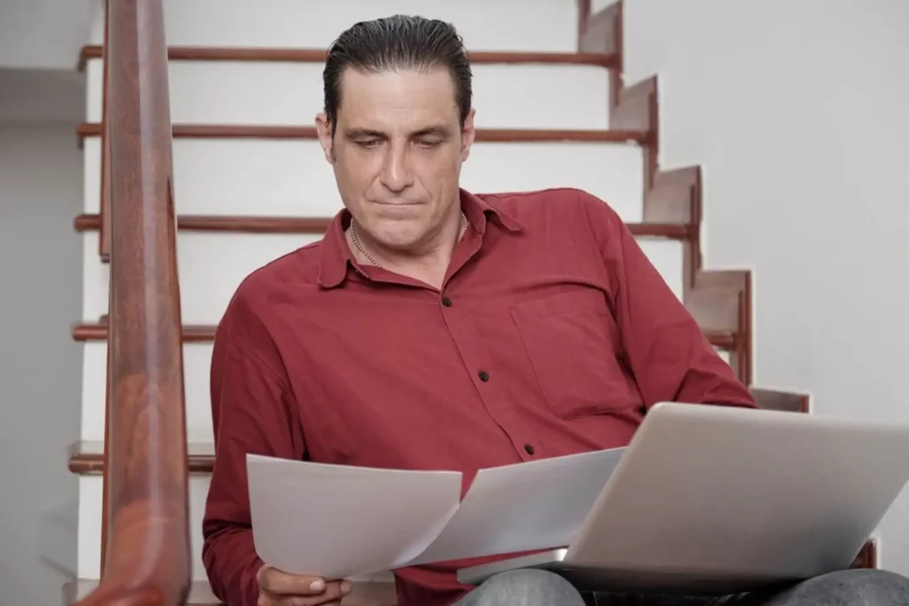 Imagem de um homem de camisa vermelha sentado na escada, olhando para os papéis que tem nas mãos, enquanto um laptop repousa em seu colo.