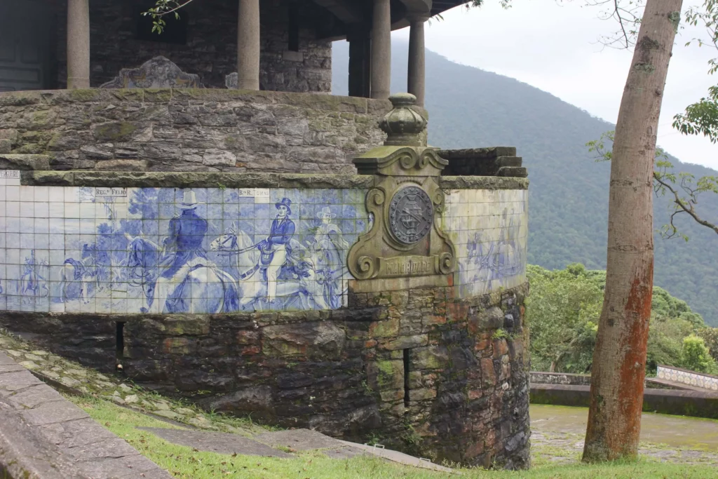 Imagem do Parque Caminhos do Mar, em São Bernardo do Campo, mostra uma estrutura de pedra com uma parede curva decorada com azulejos azuis e brancos retratando pessoas e cavalos. Uma vegetação e uma montanha aparecem ao fundo