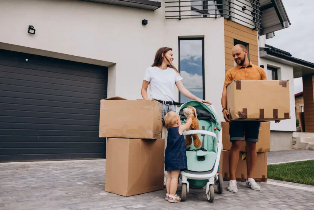 Imagem de um casal sorridente em frente a uma casa moderna, com caixas de papelão, enquanto uma criança pequena, vestindo um vestido azul, fica ao lado de um carrinho de bebê, segurando um bichinho de pelúcia.