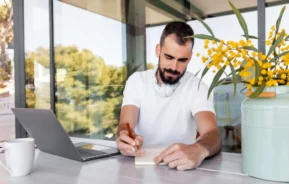 Imagem de um homem escrevendo em um bloco de notas, sentado à mesa, onde há um notebook, uma xícara de café e um vaso com flores amarelas, em uma sala moderna e bem iluminada, com paredes de vidro.
